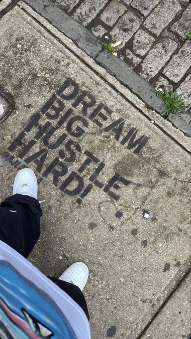 An overhead street photograph shows someone's white sneakers standing on a sidewalk next to a stenciled message on the pavement that reads 'DREAM BIG HUSTLE HARD!'