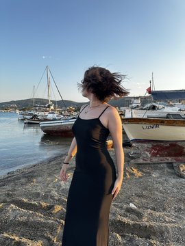 A woman in a black dress stands on rocks by a harbor with sailboats and a boat labeled 'LIZINK' in the background.