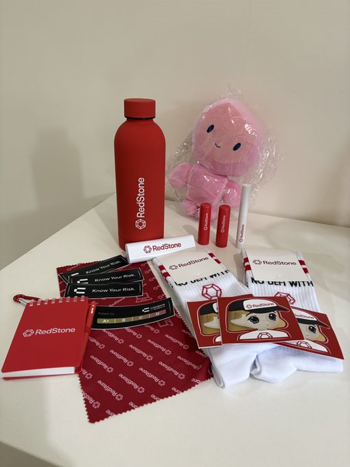 A collection of RedStone branded promotional merchandise including a red water bottle, a pink plush toy, cards, stickers, and a small notebook arranged on a white surface.