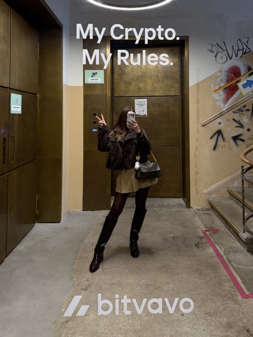 The image features a woman taking a selfie with her phone, standing in front of a door that has "My Crypt. My Rules." written above it. The background includes stairs leading up to another level of the building.