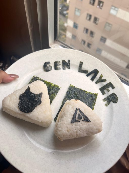A plate holding two onigiri (rice balls) decorated with seaweed cutouts in cat and Assassin's Creed shapes, with "GEN LAKER" spelled out in seaweed above them.