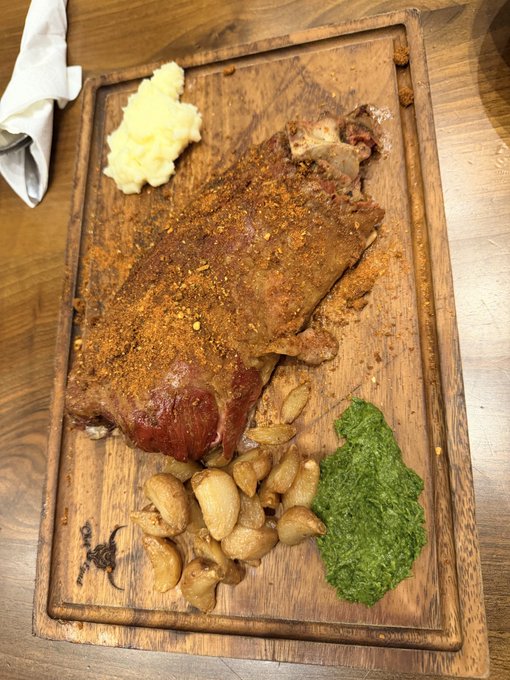 A roasted pork knuckle served on a wooden board with sides of mashed potatoes, roasted garlic cloves, and green sauce.