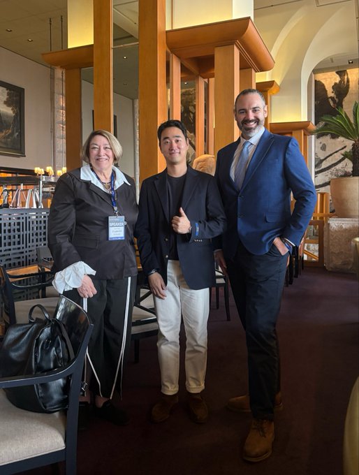 Three people pose for a photo in a hotel lobby; a woman on the left with a crutch wears a lanyard labeled 'SPEAKER', standing next to two men in blazers, one giving a thumbs-up.
