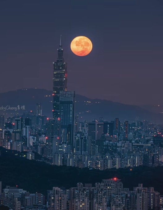 A nighttime cityscape photo credited to 'RickyCheng 影像' showing a large orange full moon rising above Taipei, with Taipei 101 tower prominently visible in the skyline.
