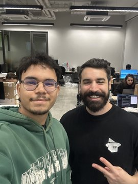 A selfie of two smiling young men in an office setting; the one on the left wears a green hoodie and glasses, while the one on the right sports a beard, black t-shirt with a monkey logo, and makes a peace sign.