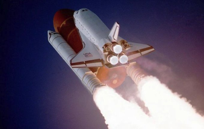 A NASA space shuttle launching into a blue sky, with its rocket boosters emitting bright flames and smoke.