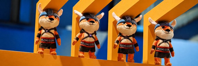 Four identical plush mascot toys wearing orange and black athletic outfits and headbands with horns, displaying 'FLOKI.COM' on their chests, are displayed in yellow alcoves.