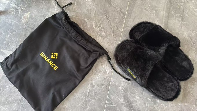Binance-branded merchandise including a black drawstring bag with the yellow Binance logo and a pair of black fuzzy slippers, displayed on a marble floor.
