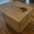 A cardboard box sealed with tape sitting on a wooden floor with a pair of scissors on top.
