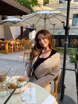 A young woman with long brown hair smiles while seated at an outdoor patio table with food and drinks, under umbrellas with stone buildings in the background.
