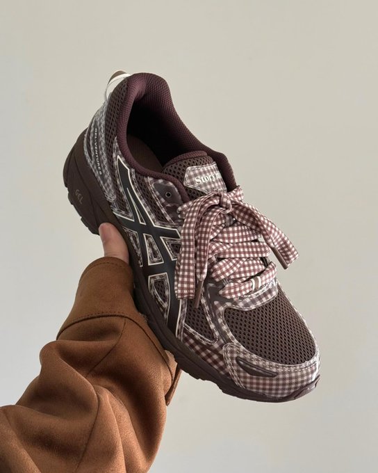 A close-up product photo of a brown and patterned Asics running shoe with gingham-print laces and ASICS branding, held by a hand in a brown sleeve against a neutral beige background.