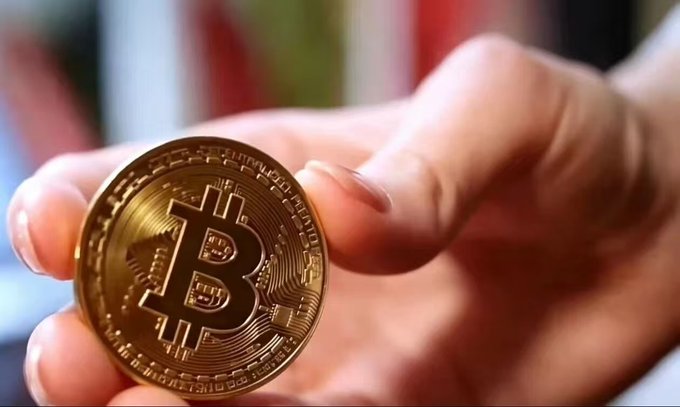 A close-up of a hand holding a physical Bitcoin token, displaying its iconic "₿" symbol with inscriptions like "BITCOIN DIGITAL DECENTRALIZED PEER TO PEER" around the edge, visually representing the focused, singular monetary purpose described in the post's comparison between Bitcoin and Ethereum.