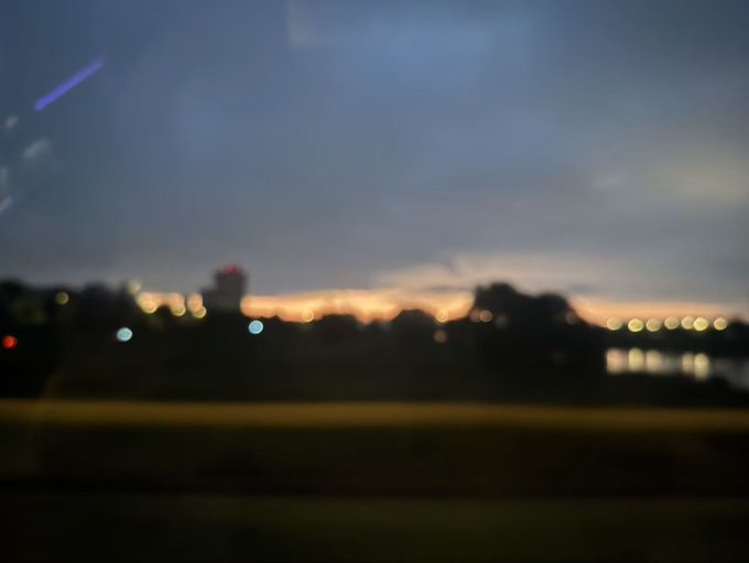 A blurry dusk skyline with distant city lights and a sunset horizon is shown, likely viewed from a moving vehicle. The image's indistinct, out-of-focus quality visually mirrors the uncertain and unclear financial situation described in the post about using a U card for RMB withdrawals.