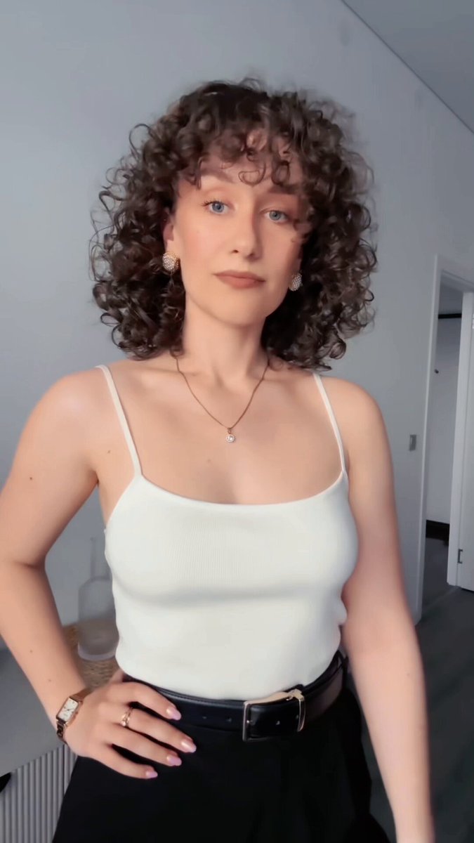 The image features a woman with curly hair wearing a white shirt and black pants. She has her hands on her hips and appears to be posing for a photo. The background of the image shows a wall, suggesting that they are indoors or in an office setting.