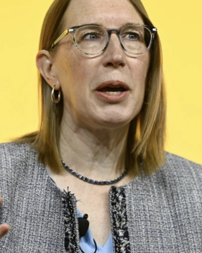 A woman with shoulder-length hair and glasses speaking, wearing a blue blazer with a black beaded necklace, photographed against a yellow background.