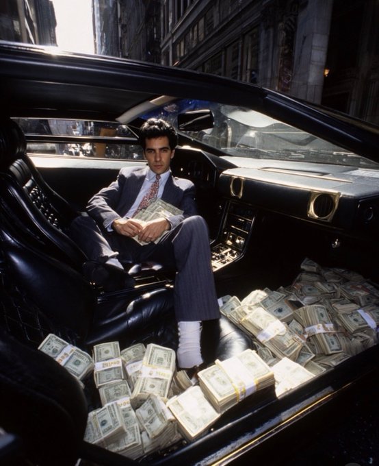 A scene from the film Scarface showing Tony Montana sitting in the driver's seat of a luxury car, wearing a suit and holding stacks of cash, surrounded by large piles of banded money on the floor.