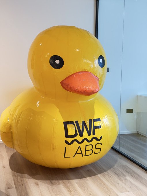 A large inflatable yellow rubber duck with "DWF LABS" printed on its side sits in an indoor space with wooden flooring.