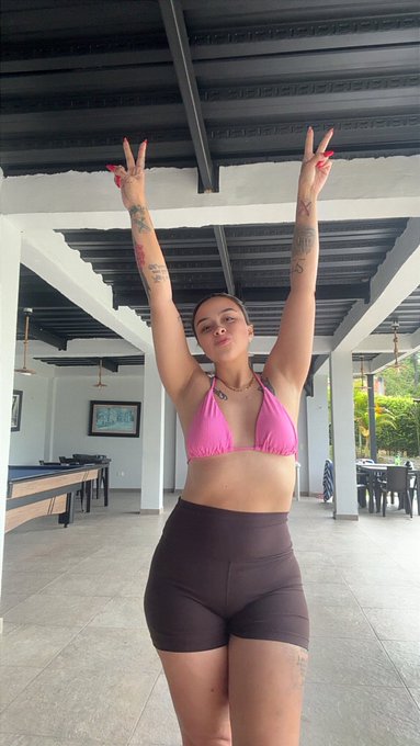 A woman in a pink bikini top and dark shorts poses with both arms raised in peace signs under a covered outdoor patio.