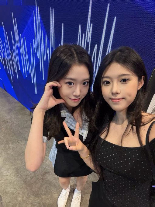 A selfie of two young women posing together indoors in front of a blue backdrop with white vertical light patterns; one is making a peace sign.