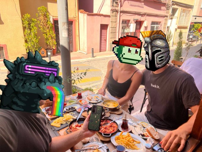 A photo of three people dining at an outdoor restaurant table full of food, with their faces covered by cartoon/video game character stickers including a mech with a rainbow, a pixel zombie, and a gladiator helmet.