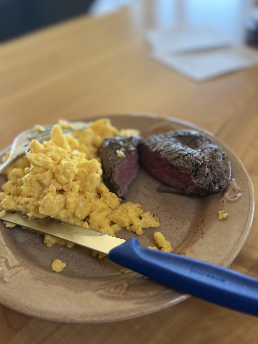 A plate of scrambled eggs and medium-rare steak slices with a blue-handled knife on a wooden table.