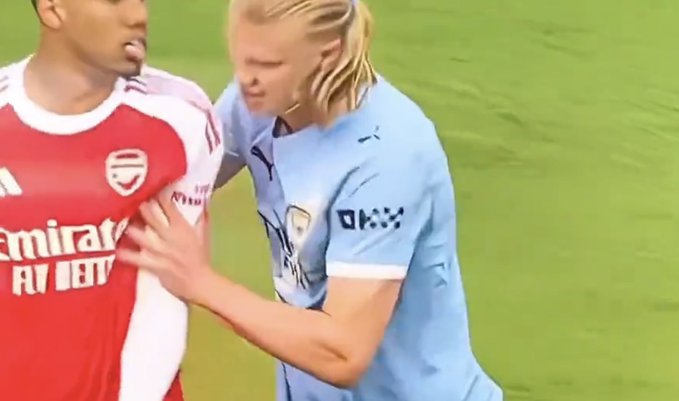 The image features two soccer players, one wearing a red jersey with "Emirates" written on it, and the other wearing a blue jersey. They are both standing on a green field, possibly during a match or practice session. The player in the red jersey is holding onto the arm of the player in the blue jersey, creating an intense moment between them.