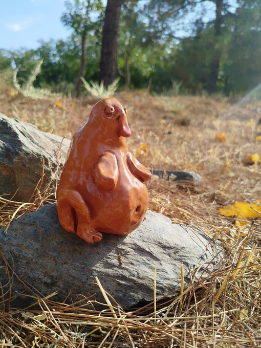 A glossy terracotta-colored figurine of a quirky creature with big lips and oversized ears sits on a rock surrounded by pine needles.
