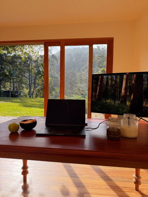 A laptop and monitor sit on a wooden table beside fruit and a candle, with large windows revealing a forest backdrop — setting the scene for a quiet home recording space to film upcoming solo SOLCAST episodes.