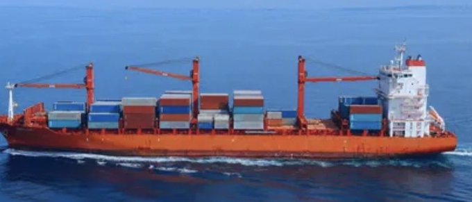 A red cargo ship loaded with multicolored shipping containers sails across the open ocean.