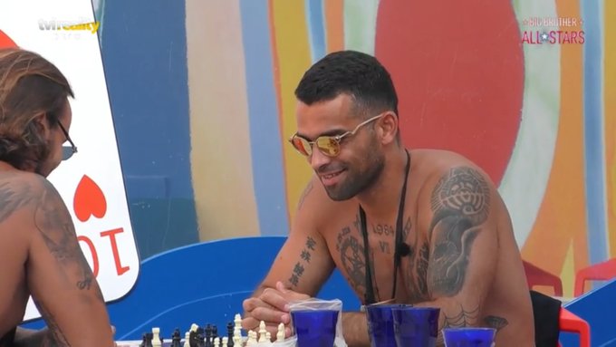 A shirtless, tattooed man wearing orange-tinted sunglasses smiles while seated at a chess board on a colorful outdoor set, with a "Big Brother All-Stars" logo visible in the upper right corner, matching the caption's playful tone of casually playing chess instead of paying attention to the show's drama.