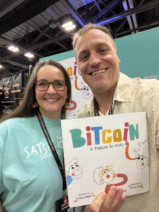 Two people proudly display a copy of the children's book "Bitcoin: A Treasure to HODL" written by Lindsey Meserow and illustrated by Kenny Menendez, with the woman wearing a teal "Sats to the Stack" shirt. The image promotes their post about donating unopened boxes of the book to classrooms, libraries, or homeschool co-ops from the Midwest Bitcoin Summit in Columbus.