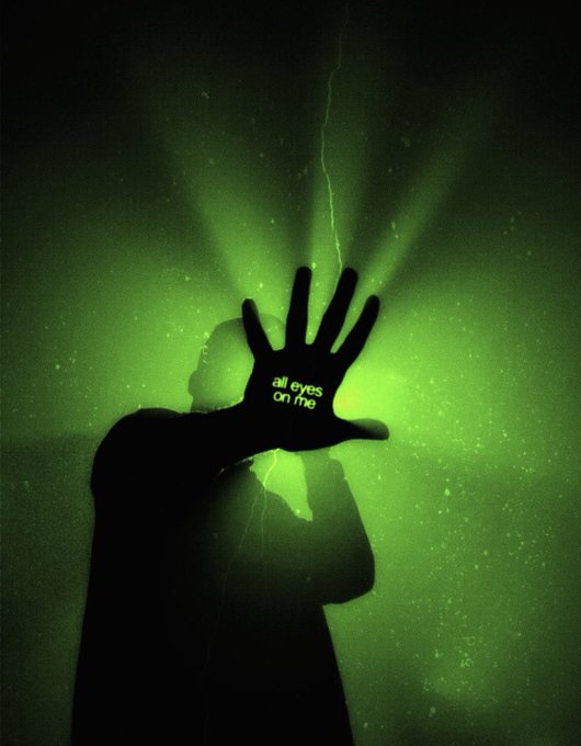 A dark, atmospheric image of a silhouetted figure with an outstretched hand displaying glowing green text 'all eyes on me,' with radiating green light beams against a black background.