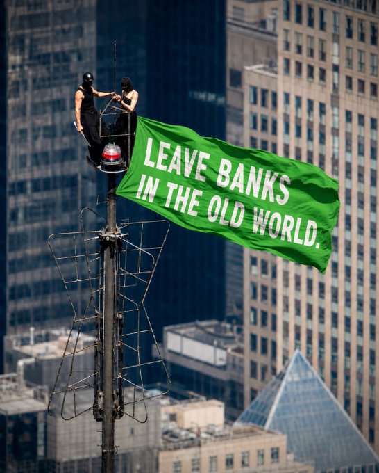 Two masked activists perched on a tall antenna display a large green banner reading 'LEAVE BANKS IN THE OLD WORLD' against a backdrop of city skyscrapers.