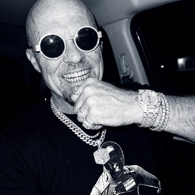 A black and white photo shows a bald man wearing round diamond-encrusted sunglasses, a large diamond chain with an astronaut pendant, and a diamond-encrusted watch.