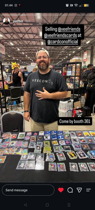 A man wearing a "VEECON" t-shirt stands behind a table covered in colorful trading cards at Card Con, with on-screen text reading "Selling @veefriends @veefriendscards at @cardconofficial" and "Come to booth 361," aligning with the post celebrating Pazbi showcasing his VeeFriends Cards collection.