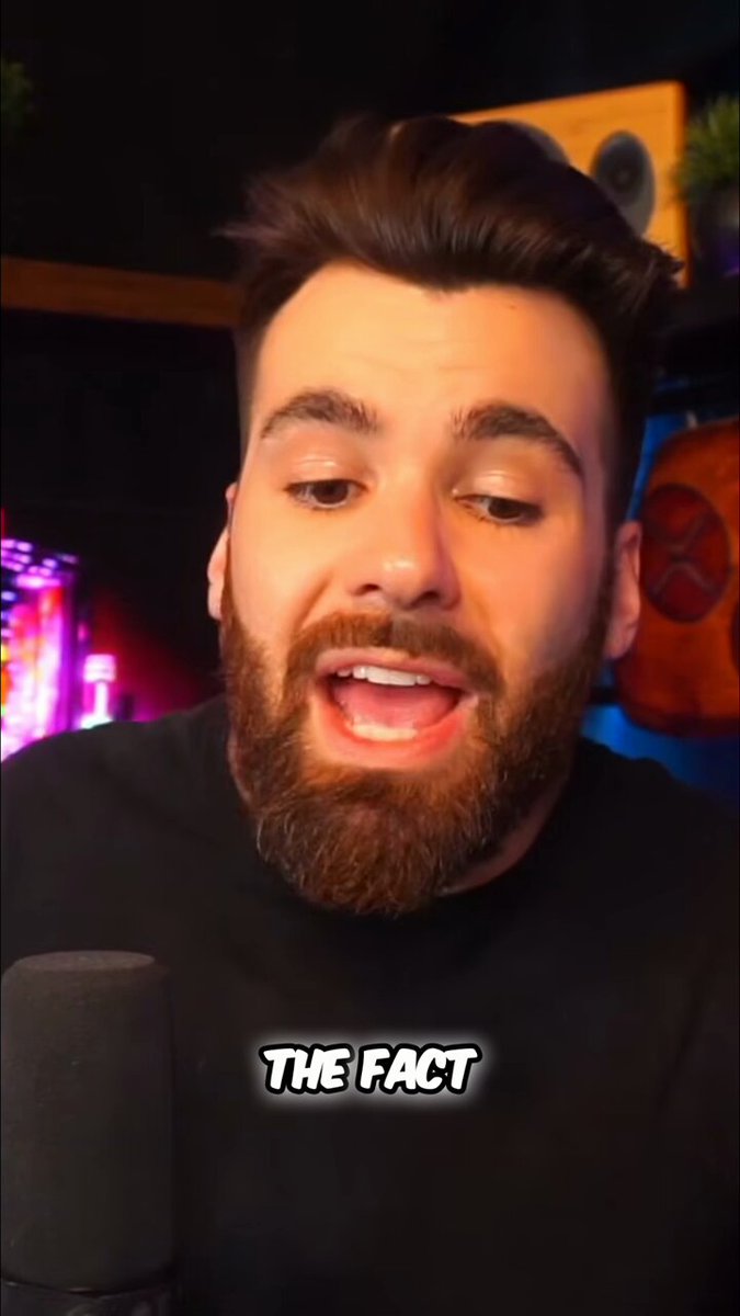 In the image, a man with a beard wearing a black shirt is speaking into a microphone while holding up a sign that says "The Market Does Not Understand the True Value of XRP #XRP #Crypto". The background features a shelf with various items on it and a plant in the corner.