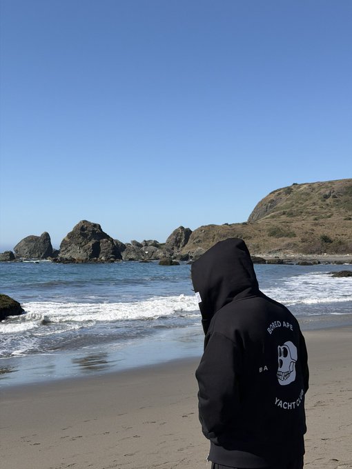 A person wearing a black hoodie with 'BORED APE YACHT CLUB' logo and skull graphic stands on a sandy beach, facing away from the camera toward rocky coastal formations and the ocean under a clear blue sky.