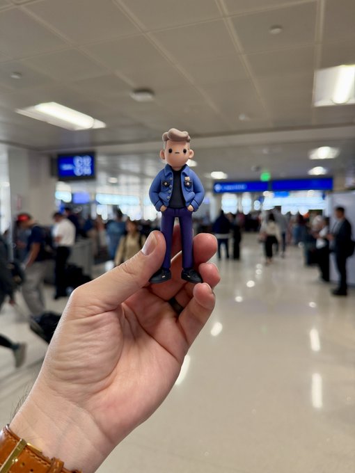 A hand holds up a small figurine of a man in a blue suit with hands in his pockets, posed in front of the blurred backdrop of an airport terminal where travelers are visible near departure boards. The playful "never fly solo" caption pairs with the scene by suggesting the toy is a tiny travel companion making the trip alongside the photographer.