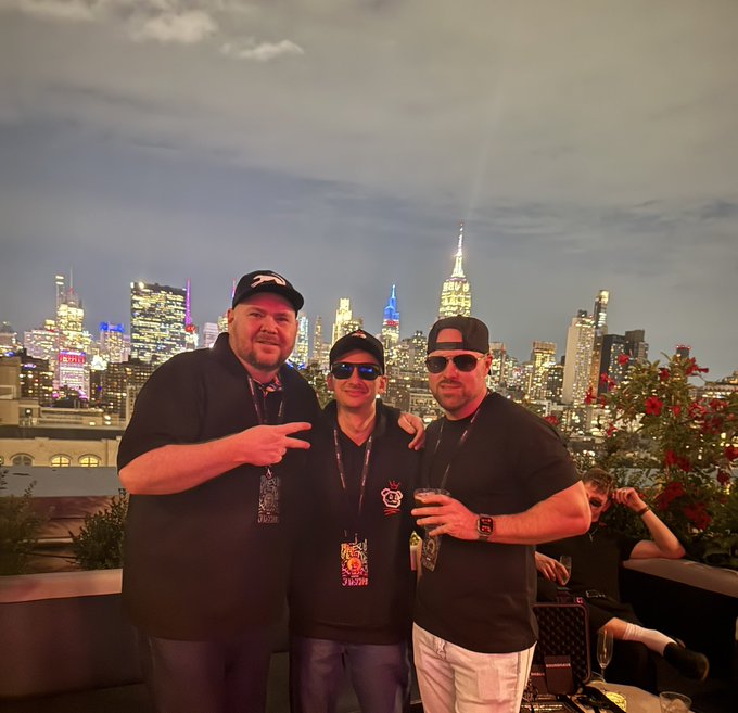 Three men wearing dark shirts and caps pose together on a rooftop at night, with the illuminated New York City skyline visible behind them.