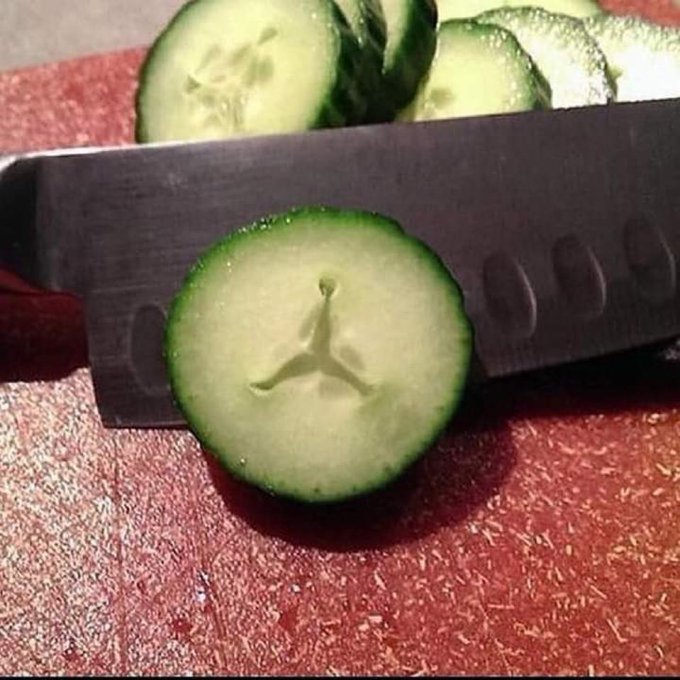 A sliced cucumber displaying a perfect Jordan logo (Jumpman silhouette) pattern in its center, positioned in front of a kitchen knife with more cucumber slices in the background.