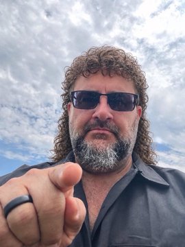 A man with curly gray-streaked hair, a beard, and sunglasses points at the camera against a cloudy sky, wearing a dark shirt and a black ring.