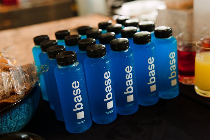 Multiple blue water bottles branded with "base" (the Base/Layer-2 blockchain) logo, displayed on a table alongside glasses of juice in a hospitality setting.