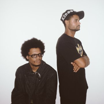 A photograph of two men standing back-to-back against a white background, one with an afro and glasses wearing a black shirt, and the other wearing a black cap and black t-shirt with crossed arms.