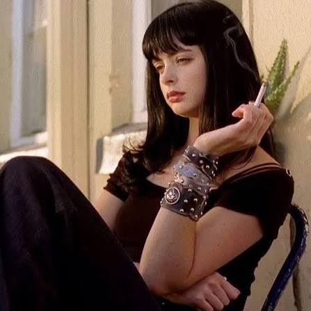 A young woman with dark hair and bangs, wearing multiple studded bracelets and smoking a cigarette, sits in a dimly lit room.