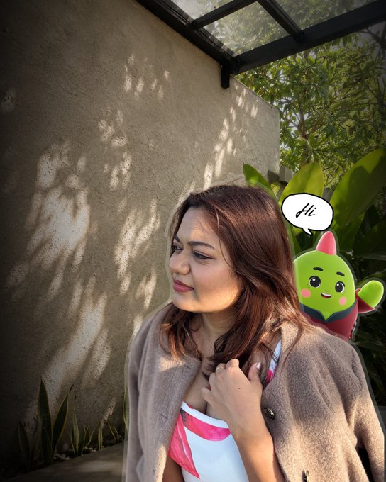 The image features a woman standing next to a wall, wearing a brown coat. She has her arm around another character from the game, which appears to be a green alien or cactus with a face. The character seems to be waving at the woman as if they are having a conversation. The background of the image is filled with trees and plants, creating an outdoor setting for this interaction between the woman and the character in the game.