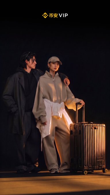 Two people pose together in a stylized photoshoot with dramatic lighting, standing next to a suitcase, with '币安VIP' (Binance VIP) text and logo at the top.