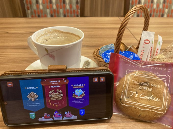 A smartphone playing a mobile game (showing 'Season 18' Casual/Ranked/Arcade modes) sits on a table next to a cappuccino, a cookie labeled 'TAKAKURA MACHI COFFEE T's Cookie', and a basket of condiments.