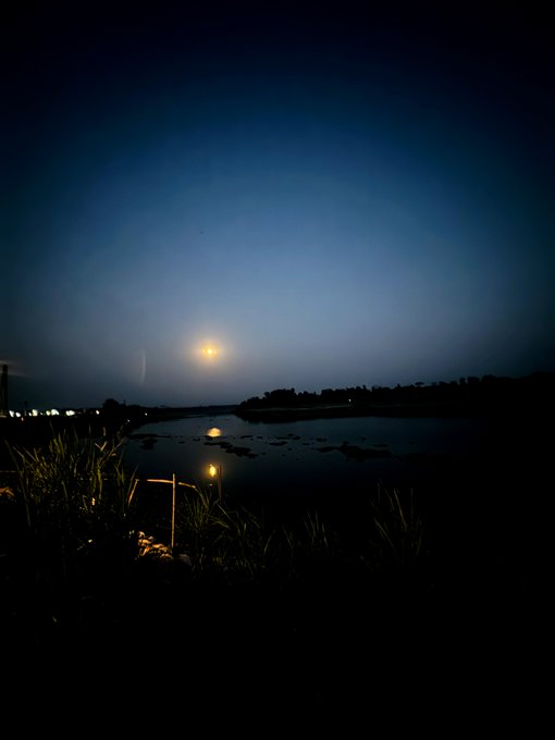 A nighttime photograph of a bright full moon reflecting over a calm body of water, with silhouetted grass in the foreground and distant city lights on the horizon.