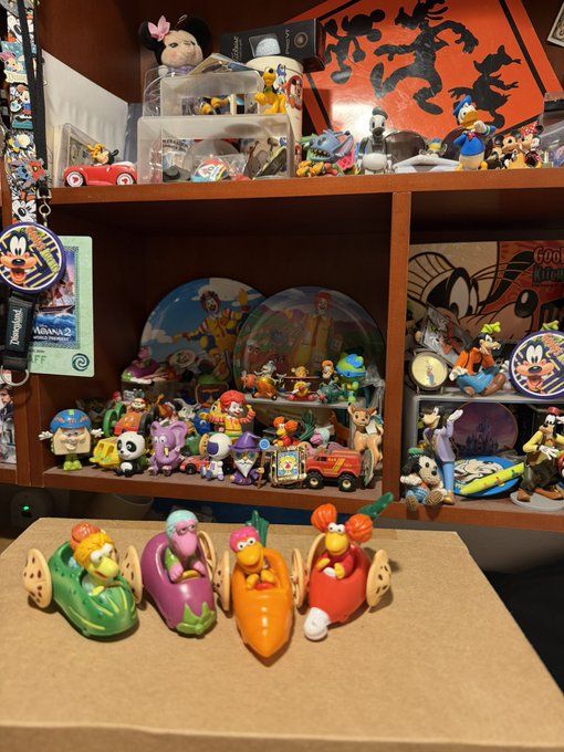 A collection of Disney-themed toys and figurines is displayed on wooden shelves and on cardboard boxes in the foreground, including characters from the Muppets, Donald Duck, and others.