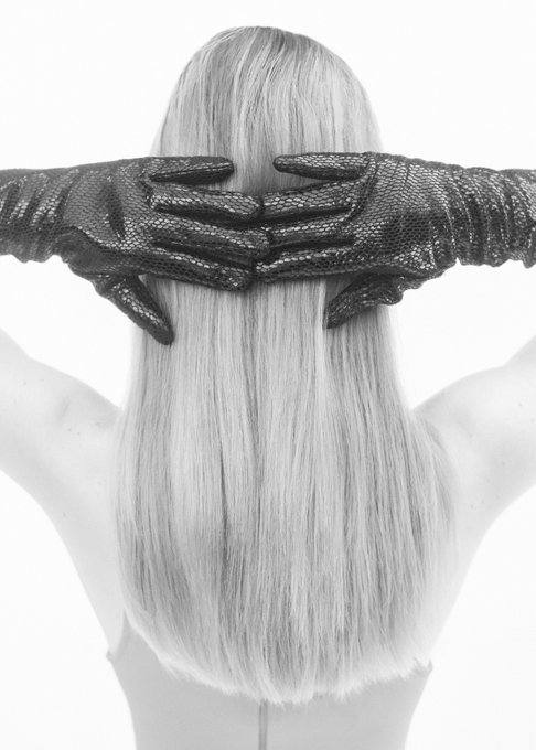 A black-and-white artistic photograph showing a person from behind with long hair, their face covered by gloved hands.